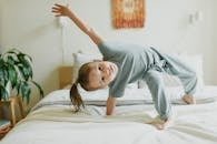 A playful young girl in gray pajamas enjoys a moment of fun on a bed in a cozy bedroom setting.